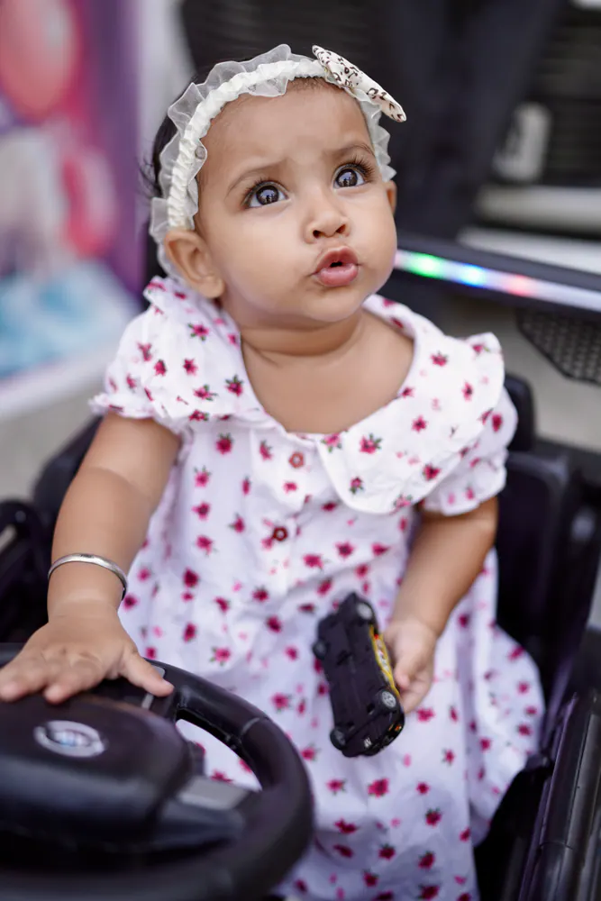 Baby girl in a polka-dot frock sitting at the wheel of a toy car