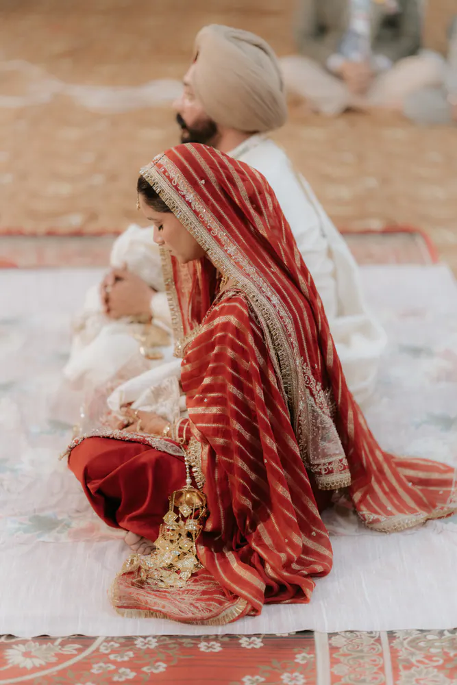 Sikh bride in a red striped dupatta seated beside the groom during the wedding rituals